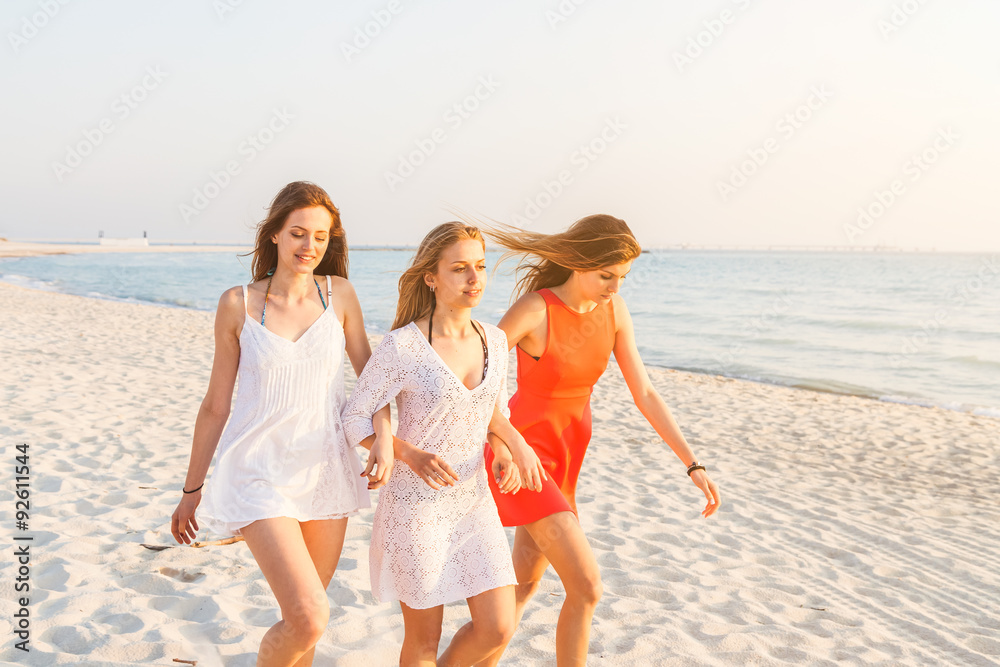 After a day at sea, a group of three young women walking barefoot on the sand in the rays of the setting sun. Behind them the deserted beach without people