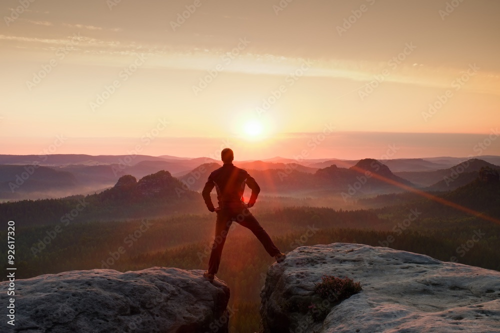 Obraz premium Jumping hiker in black celebrate triumph between two rocky peaks. Wonderful daybreak with sun above head..