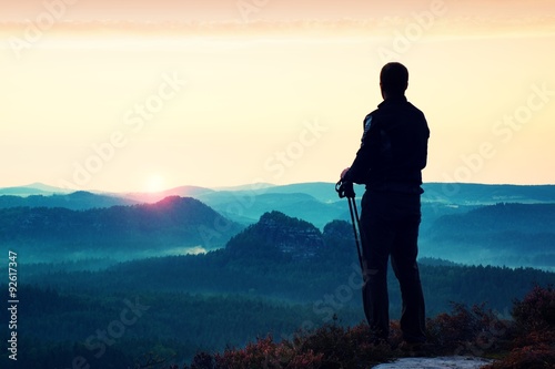 Silhouette of tourist guide with poles in hand. Hiker with sporty sportswear stand on sharp view point above misty valley. Sunny daybreak in rocky mountains.