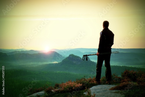 Silhouette of tourist guide with poles in hand. Hiker with sporty sportswear stand on sharp view point above misty valley. Sunny daybreak in rocky mountains. photo