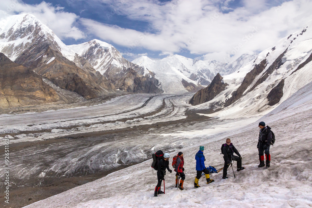 Group of Hikers Walking on Snow and Ice Terrain Large Group of People Sport Clothing Climbing Gear High Altitude Boots Going Up Mountain Peaks Sunlight Cloud Sky Massive Glacier Flow on Background