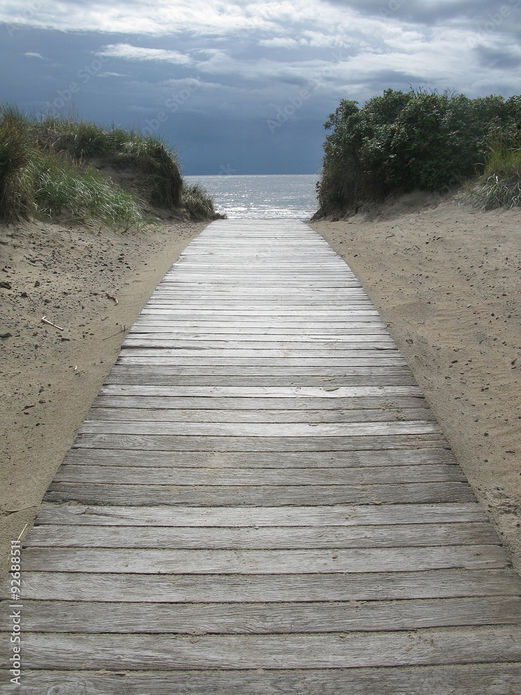 Sand dune landscape with wooden boardwalk leading towards sea and sky at Skrea Strand on a sunny day with dark clouds in Falkenberg, Sweden.