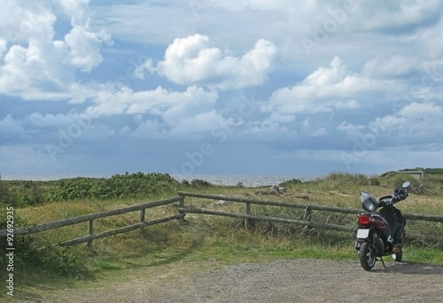 Motorcycle parked on gravel road by wooden fence in natural heath landscape at Skrea Strand on a sunny day with dark clouds in Falkenberg, Sweden. © artesiawells