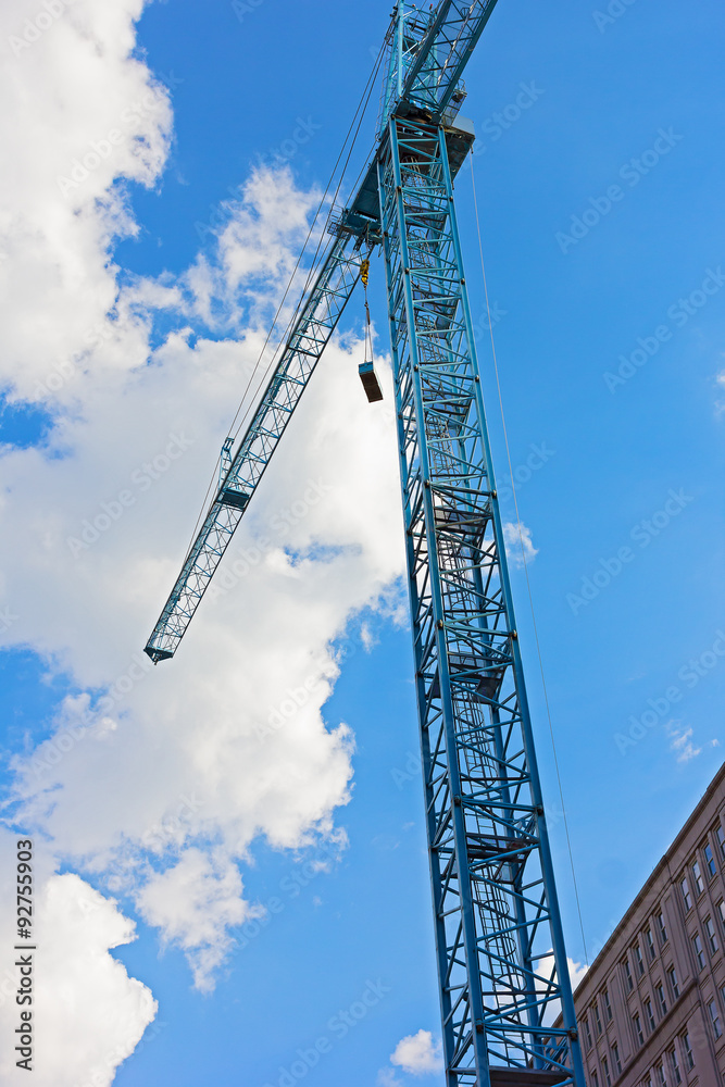 Blue construction crane against a cloudy blue sky. Construction crane lifts a cargo at urban development site. Blue construction crane against a cloudy blue sky. Construction crane lifts a cargo at urban development site.