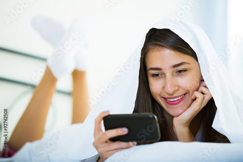 Smiling girl with smartphone hiding in bed photo