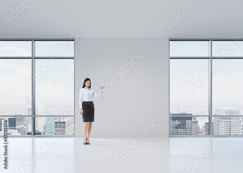 Full length brunette girl in a formal clothes is pointing out something by her hand on the white wall. A modern panoramic office in Manhattan, New York.
