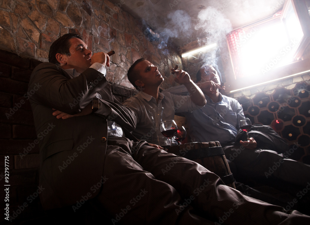 Three businessmen sitting in the wine cellar drinking wine and smoke cigar, resting after a hard day at work.