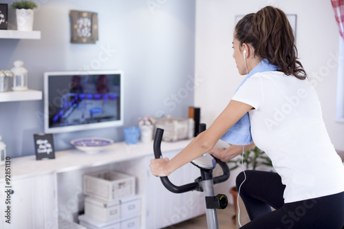 Young sporty woman training on exercise bike in the living room  and watching tv