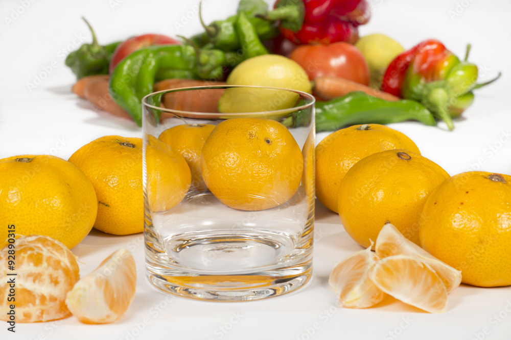 Empty glass cup next to several mandarins and tangerine segments. A bunch of different vegetables at background Empty glass cup next to several mandarins and tangerine segments. A bunch of different vegetables at background