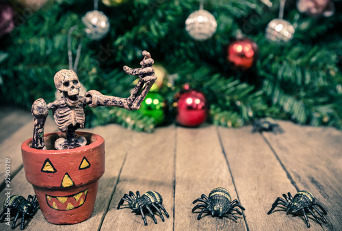 Obraz na plátne Funny Skeleton Sitting in Halloween Pumpkin Pot and Tiny Spiders on Wooden Backg