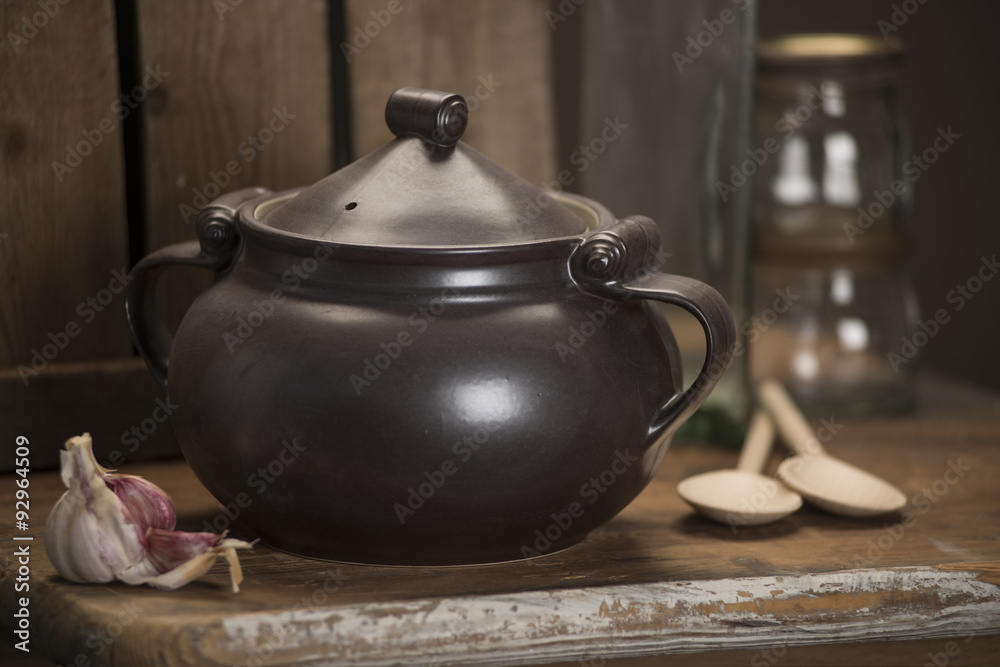 Covered black tureen alongside cloves of garlic and a pair of wooden spoons on a wooden counter.