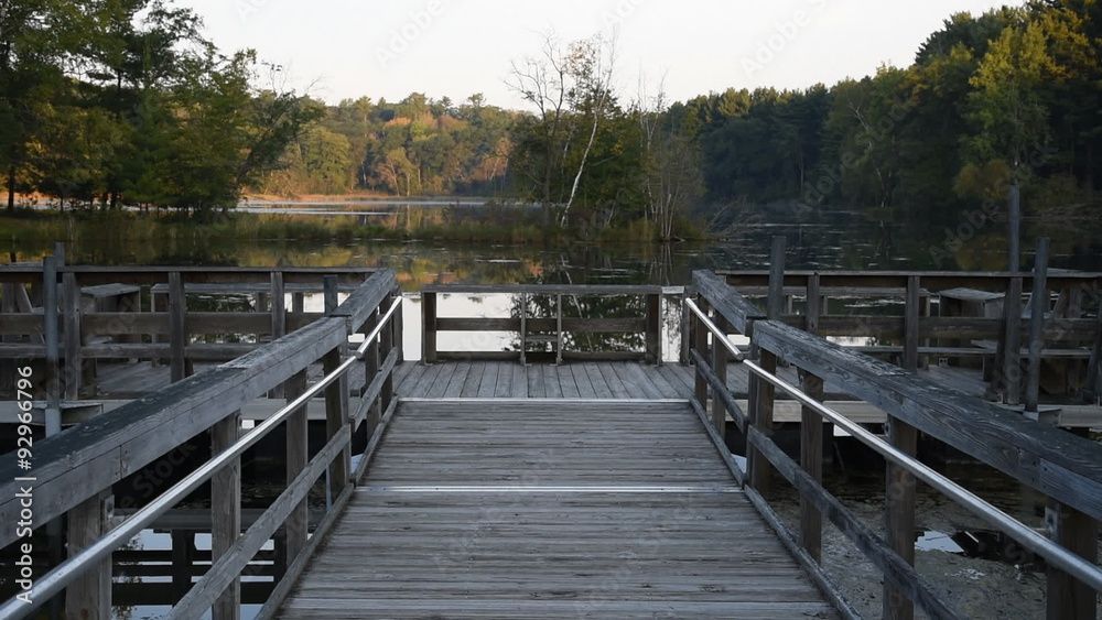 A floating wooden fishing dock sits on the calm waters of pond surrounded by trees, in the early morning. A floating wooden fishing dock sits on the calm waters of pond surrounded by trees, in the early morning.