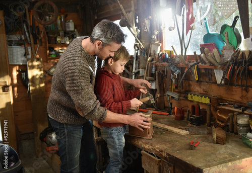 Father and his son working together in a wooden workshop. They are focused on building a wooden bird house, the son is knocking with a hammer while his grey hair father is holding the house © jackfrog