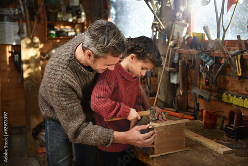 Portrait of a middle age gray hair man with beard working with his little girl in the workshop. They are focused while hammering a nail on a wooden birdhouse. They are wearing jeans and woolen pulls. © jackfrog