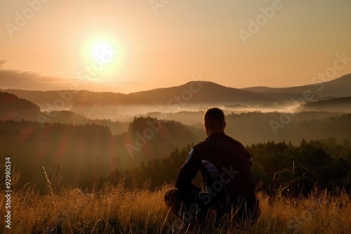 Hiker in squatting position in high grass meadow enjoy the colorful sunrise scenery