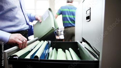 A male office worker placing a folder inside a filing cabinet before closing the drawer and leaving the scene.