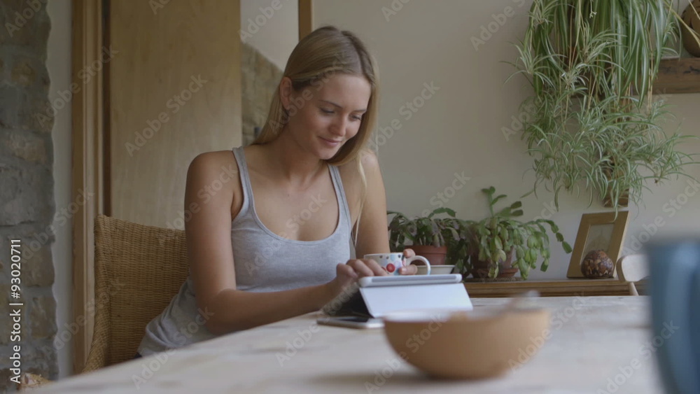 A young woman is sitting in her home enjoying a cup of tea whilst using social media on her digital tablet. A young woman is sitting in her home enjoying a cup of tea whilst using social media on her digital tablet.