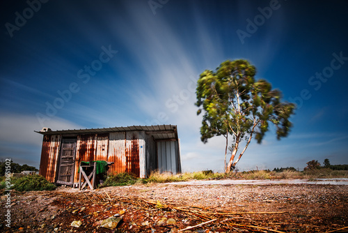 Fototapet Old shack made of sheet metal next to a tree with a long exposure