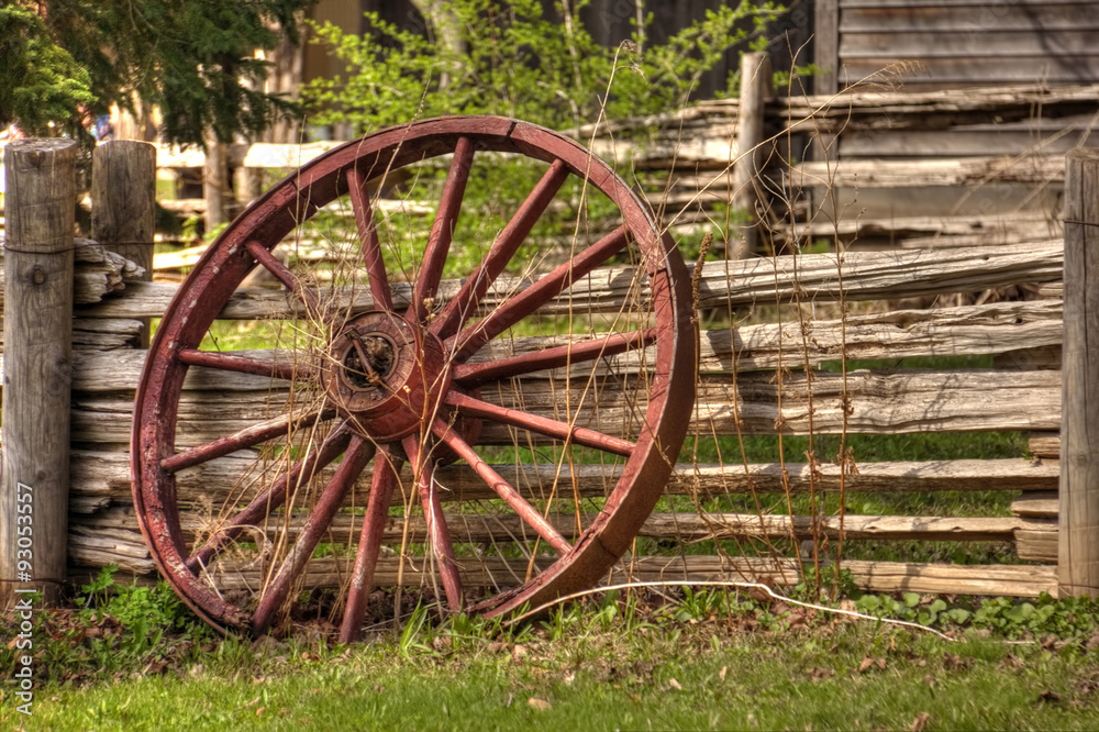 A single rustic wagon wheel leaning against wooden fence.