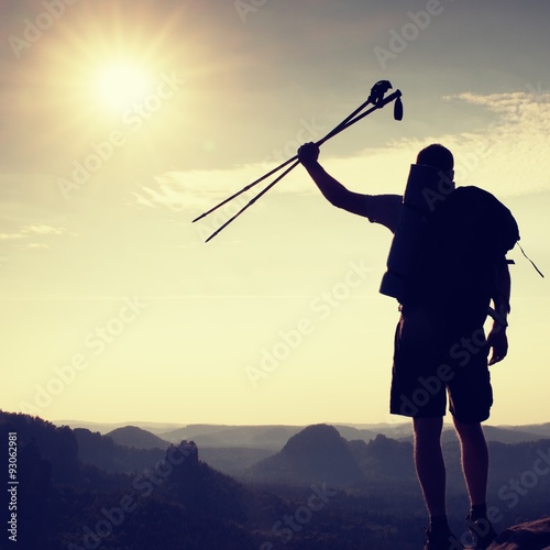 Silhouette of tourist with poles in hand. Sunny daybreak in rocky mountains. Hiker with sporty backpack, view point above misty valley.