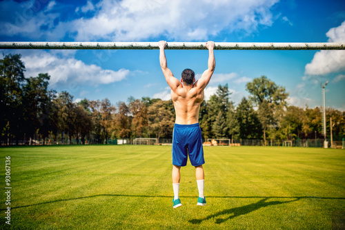 athletic built man doing chin-ups and core training in park. Fitness football player training on grass court, hardcore training