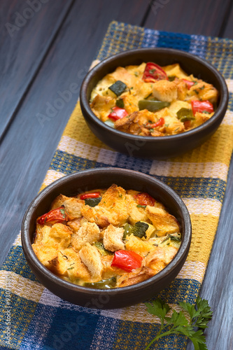 Savory baked vegetarian bread pudding made of zucchini, bell pepper, tomato and diced baguette, photographed on dark wood with natural light (Selective Focus, Focus in the middle of the first dish)