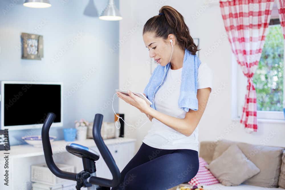 Sporty woman training on exercise bike using tablet in bright living room