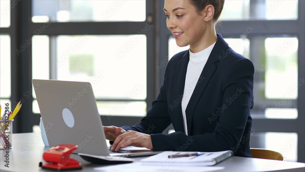 Tilt up slow motion shot of female professional using laptop. Smiling young businesswoman typing on laptop keyboard. She is sitting at desk in brightly lit office. Tilt up slow motion shot of female professional using laptop. Smiling young businesswoman typing on laptop keyboard. She is sitting at desk in brightly lit office.