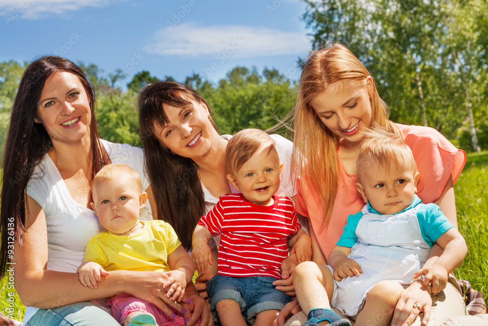 Three women with their babies sit on green grass