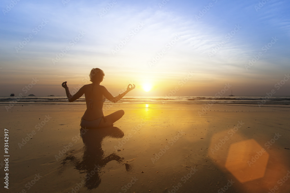 Beautiful girl sitting on beach during sunset and meditating in yoga pose.