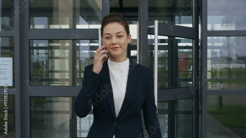 Slow motion video of beautiful businesswoman opening doors while leaving from office. Confident professional using smart phone. She is walking outside office building.