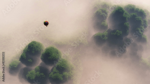 Aerial of hot air balloon floating above misty forest.