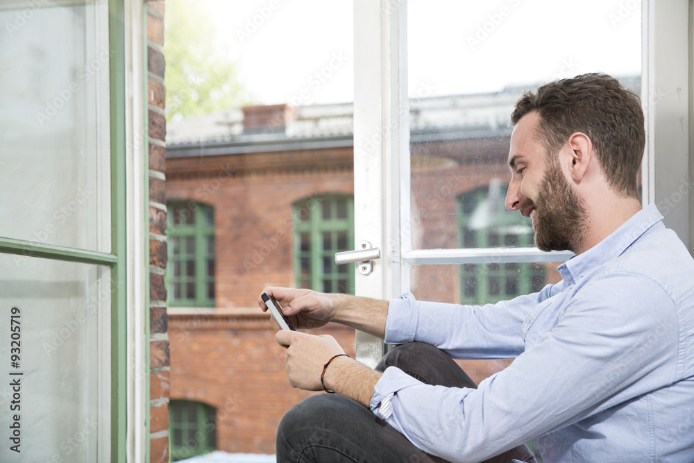 Smiling young man with cell phone sitting at the window Smiling young man with cell phone sitting at the window