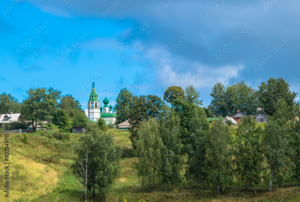 dark skies rolling in over community of Russian homes and white church with bell tower with green roof and domes 
