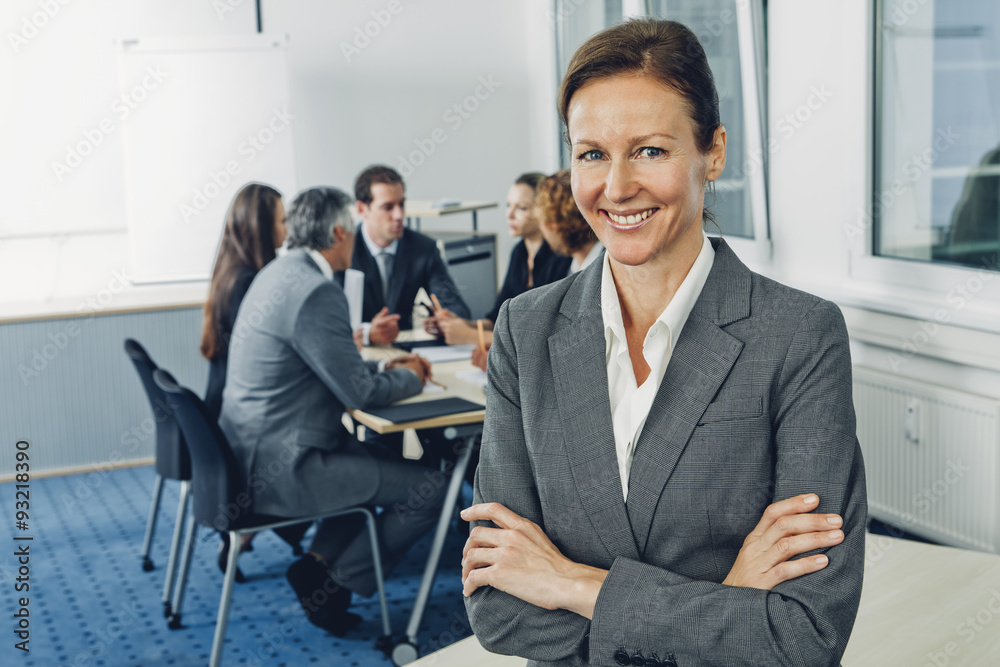 Mature businesswoman sitting on desk with arms crossed, team working in background Mature businesswoman sitting on desk with arms crossed, team working in background