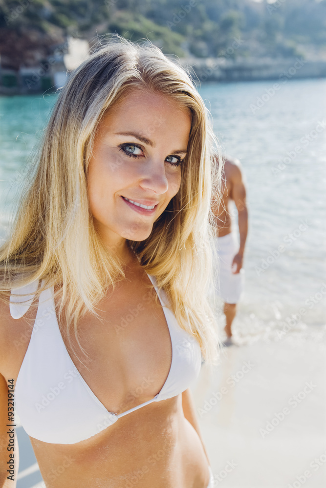 Spain, Majorca, smiling woman in bikini on the beach with man in background Spain, Majorca, smiling woman in bikini on the beach with man in background
