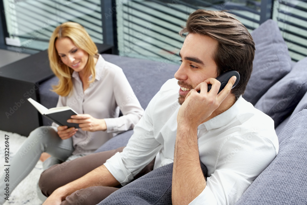 Smiling young man talking on telephone while his girlfriend sitting in the background reading a book Smiling young man talking on telephone while his girlfriend sitting in the background reading a book