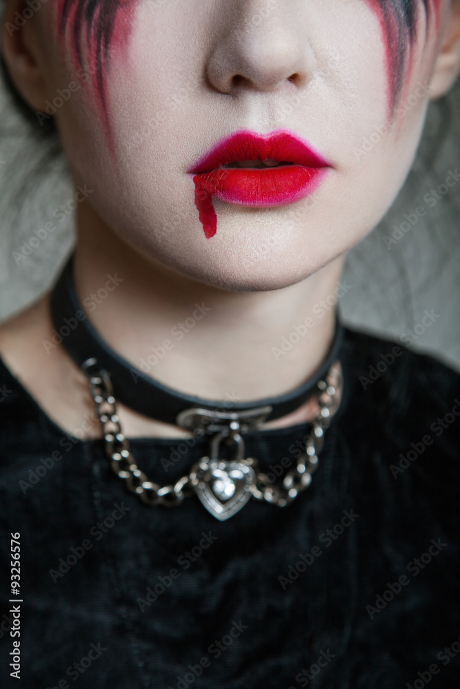 Young beautiful gothic woman with white skin and red lips with bloody drops wearing black collar with spikes. Red smokey eyes. Halloween makeup.