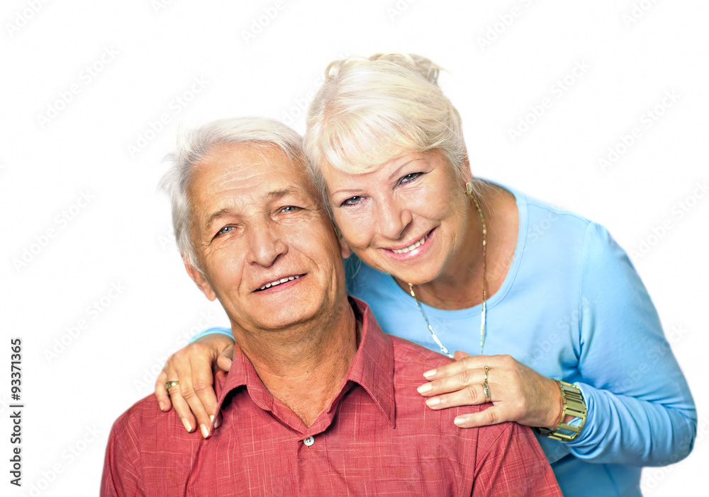 Seniorin steht hinter ihrem sitzenden Mann und umarmt ihn von hinten, beide lächeln in die Kamera Seniorin steht hinter ihrem sitzenden Mann und umarmt ihn von hinten, beide lächeln in die Kamera