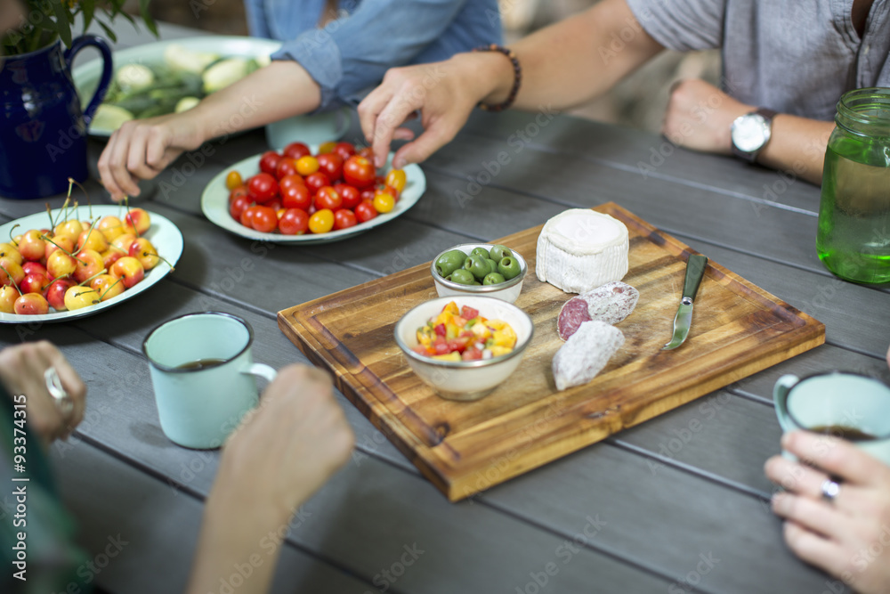 A group of people gathered around a table with plates of fresh fruits and vegetables, and a round cheese and salami on a chopping board. A group of people gathered around a table with plates of fresh fruits and vegetables, and a round cheese and salami on a chopping board.