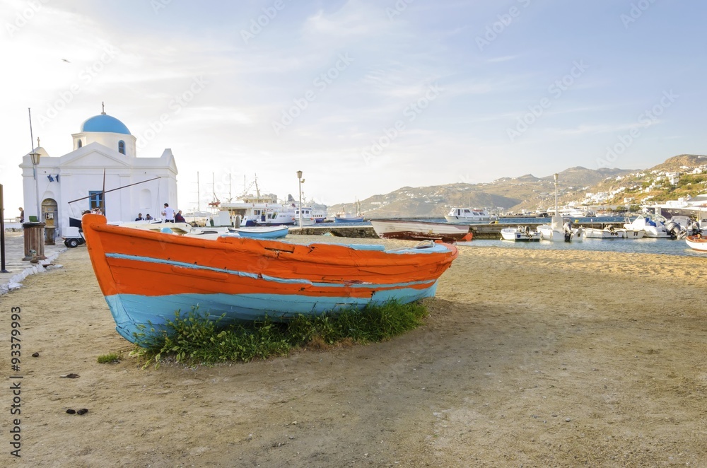 Fototapeta premium Colourful fishing and sail boats anchored on the Chora port in Mykonos,Greece. View of orange,blue boat on sand shore and the whitewashed blue dome church of Hora harbour, a greek island landscape.