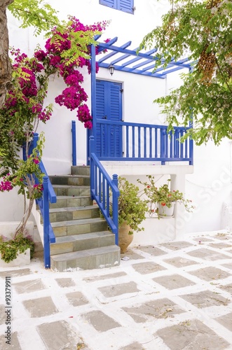 A very traditional alley view of the architecture in Chora,on the greek island Mykonos,Greece.A blue door,fence,windows,red bougainvillea, flowers outside a whitewashed house and cobble paved street
