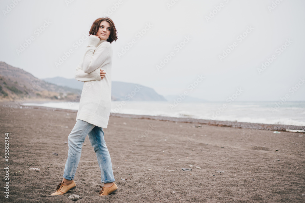 Beautiful young woman wearing white sweater and blue jeans walking on a lonely beach in a cold windy weather