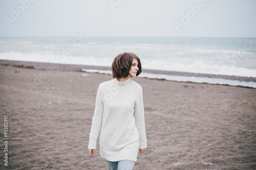 Beautiful young woman wearing white sweater and blue jeans walking on a lonely beach in a cold windy weather