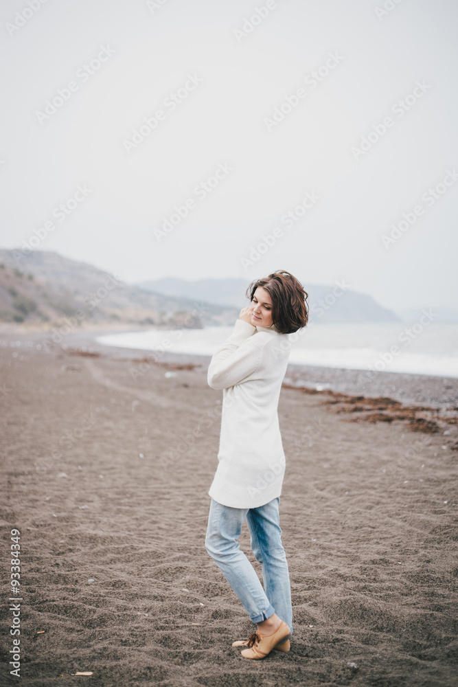 Beautiful young woman wearing white sweater and blue jeans walking on a lonely beach in a cold windy weather