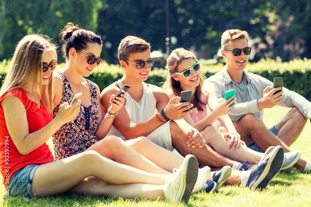 smiling friends with smartphones sitting on grass smiling friends with smartphones sitting on grass