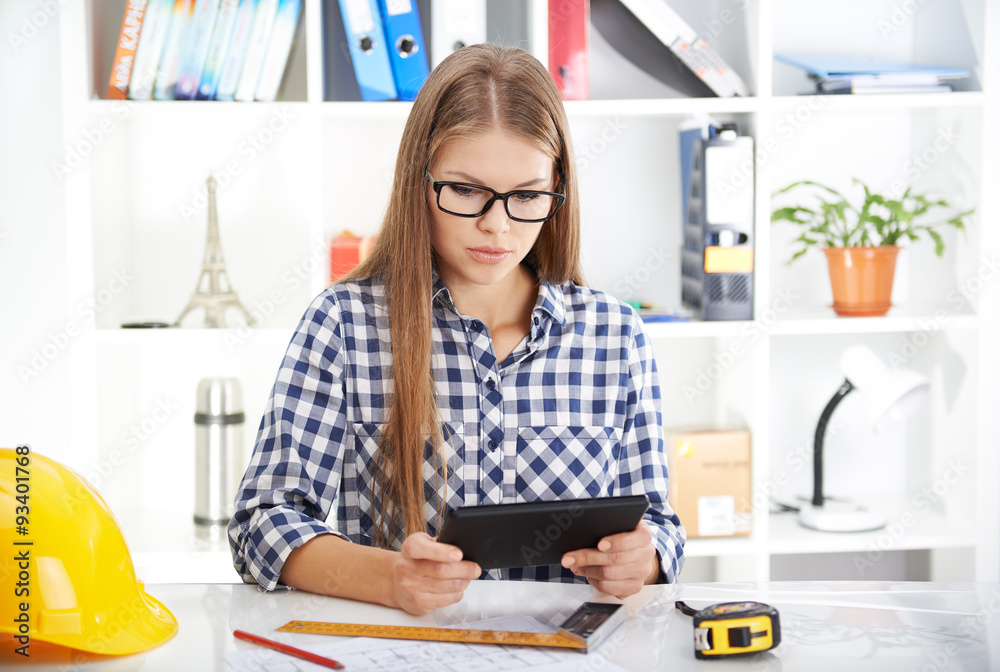 Concentrated female architect with digital tablet studying construction plans. Young woman designer sitting at table with computer and engineering tools. Concentrated female architect with digital tablet studying construction plans. Young woman designer sitting at table with computer and engineering tools.