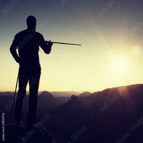 Tourist guide show the right way with pole in hand. Hiker with sporty backpack stand on rocky view point above misty valley. Sunny spring daybreak in rocky mountains.