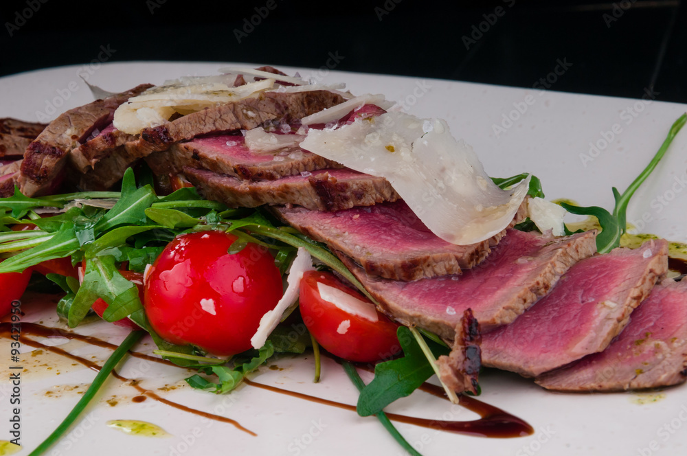 Medium rare steak served on a plate with cherry tomatoes and arugula. On top of it is grated parmesan cheese. Medium rare steak served on a plate with cherry tomatoes and arugula. On top of it is grated parmesan cheese.