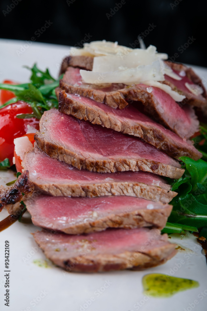 Medium rare steak served on a plate with cherry tomatoes and arugula. On top of it is grated parmesan cheese. Medium rare steak served on a plate with cherry tomatoes and arugula. On top of it is grated parmesan cheese.